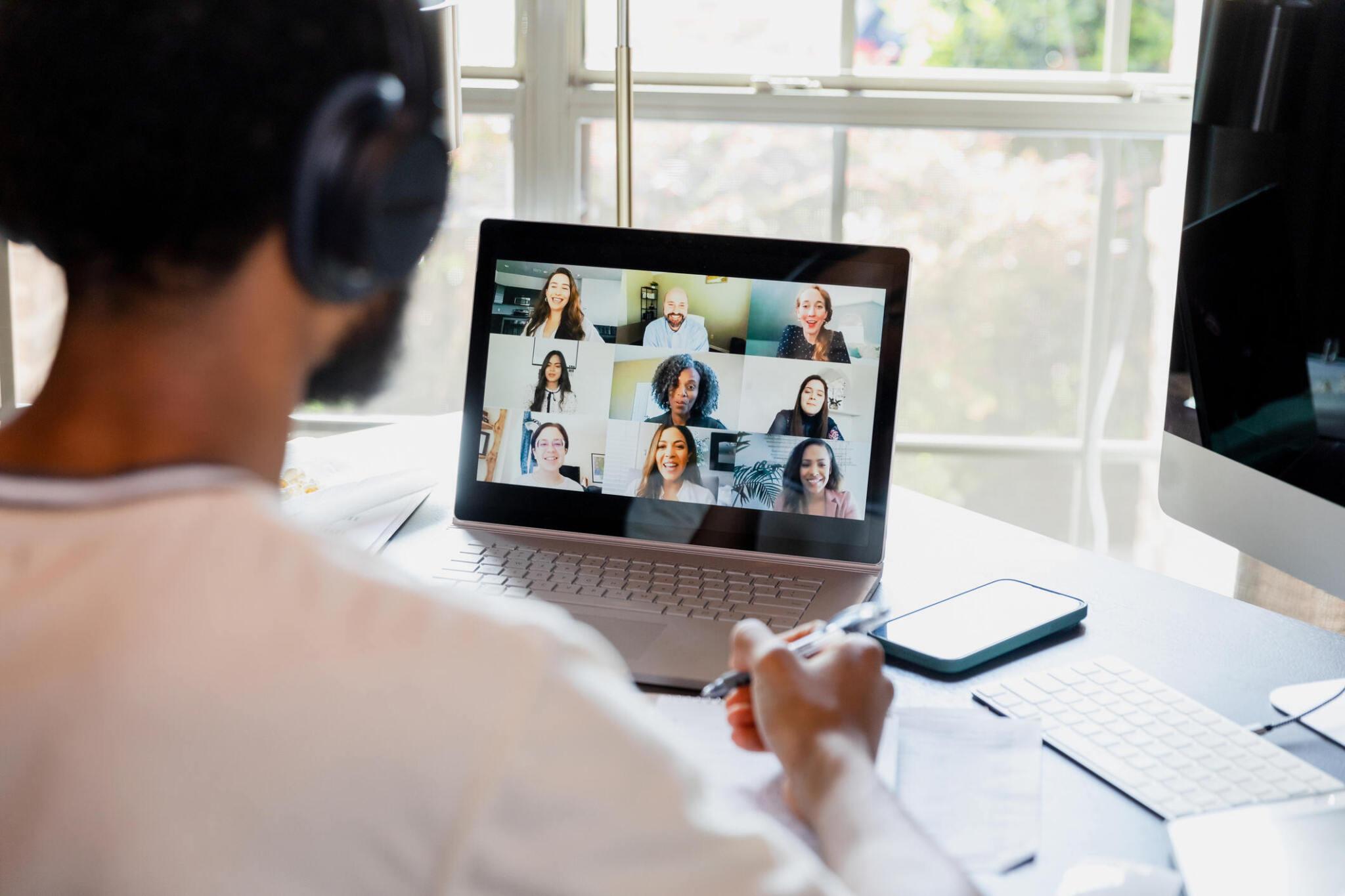 An over-the-shoulder view of an university student meeting online with his teacher and fellow students through his laptop computer and headphones.