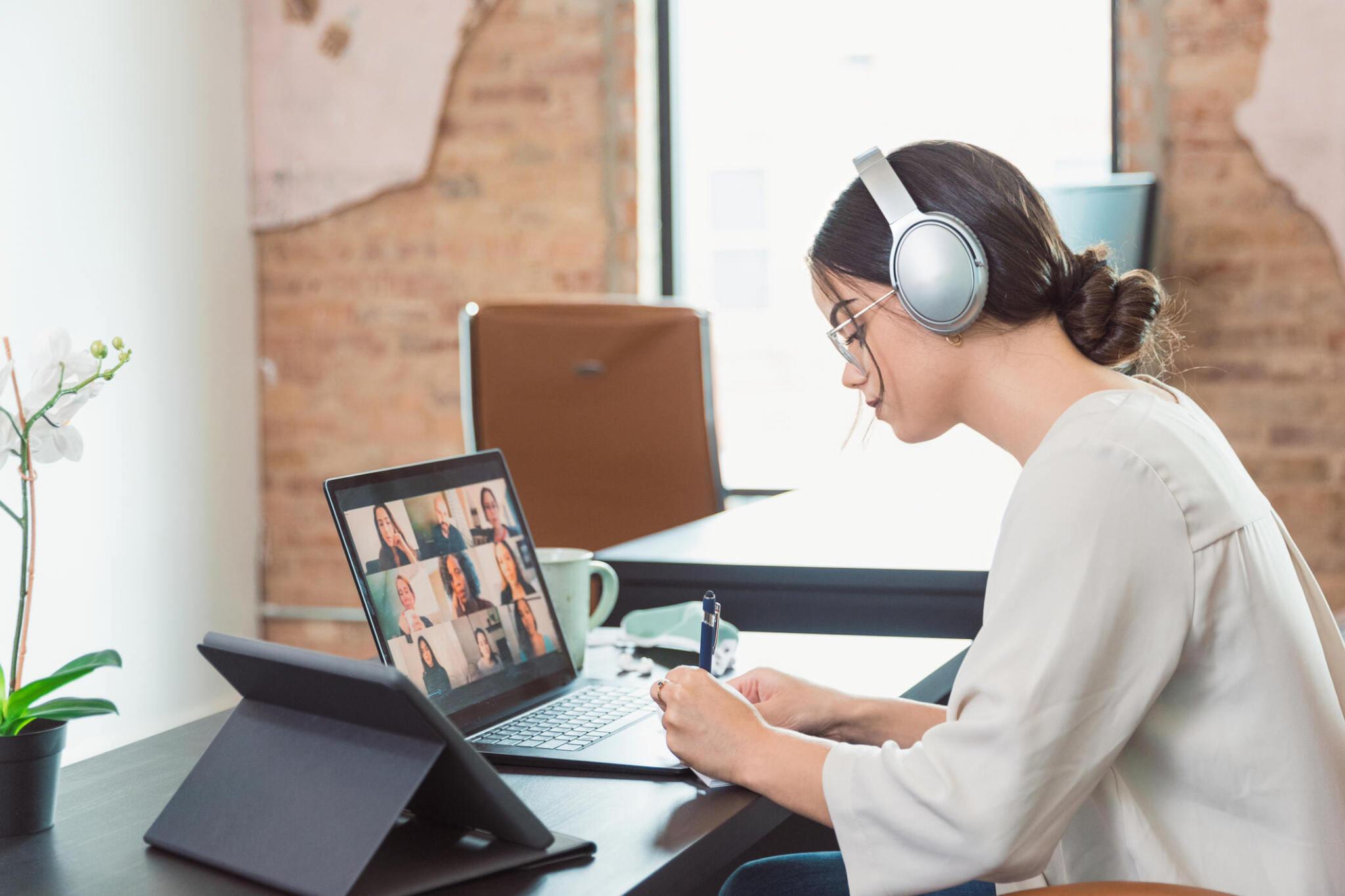 Business woman takes notes during virtual meeting with diverse colleagues using bose qc wireless headphones and laptop