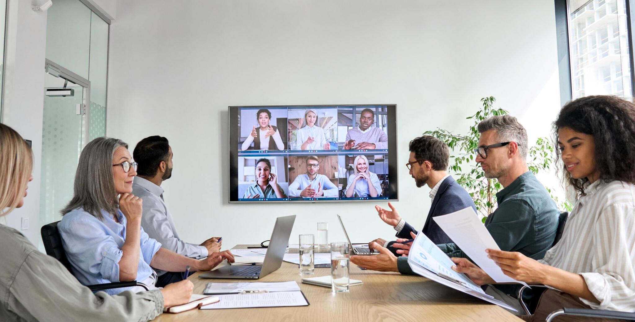 Some people are sitting in the office looking at the big display while discussing with other colleagues in online meetings