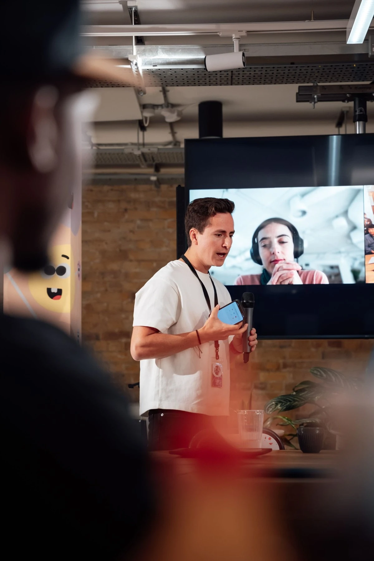 A presenter speaking in a room while a remote colleague with a headset joins on the big screen